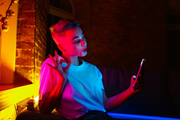 Miły. Kinowy portret stylowej kobiety w oświetlonym neonem wnętrzu. Stonowane jak efekty kinowe, jasne neonowane kolory. Kaukaski model za pomocą smartfona w kolorowe światła w pomieszczeniu. Kultura młodzieżowa.