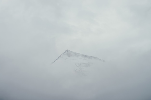 Minimalistyczny górski krajobraz z wysoką spiczastą skałą w chmurach. minimalistyczna górska sceneria z ostrym ośnieżonym szczytem górskim nad chmurami. śnieżnobiały spiczasty szczyt nad białymi chmurami. duży top w gęstej mgle