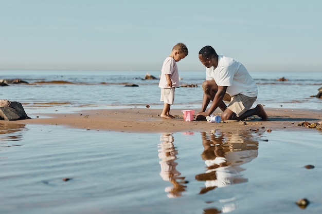 Minimalistyczny widok z boku portret młodego afrykańskiego ojca i syna bawiących się piaskiem na plaży razem...