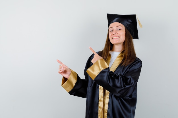 Młoda absolwentka wskazując w lewym górnym rogu w akademickim stroju i patrząc energicznie. przedni widok.