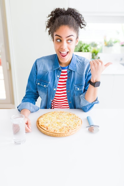 Młoda Afroamerykanka jedząca domową pizzę z serem, wskazująca i pokazująca kciukiem do boku z uśmiechniętą szczęśliwą twarzą