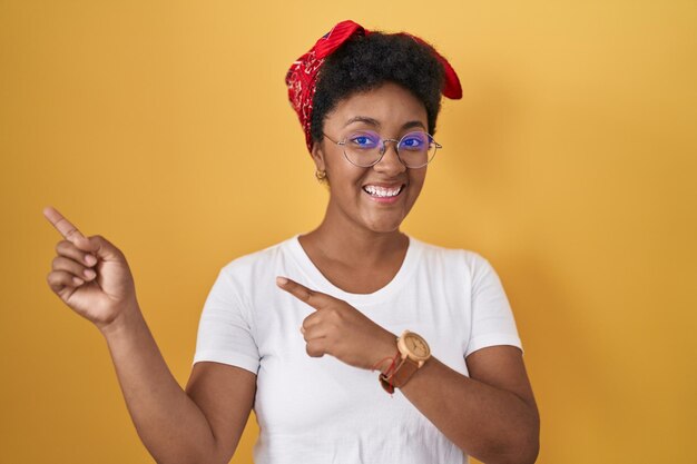 Młoda Afroamerykanka stojąca na żółtym tle uśmiechająca się i patrząca w kamerę wskazującą dwiema rękami i palcami na bok