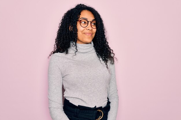 Młoda Afroamerykanka ubrana w sweter z golfem i okulary na różowym tle uśmiecha się patrząc w bok i odwracając wzrok myśląc