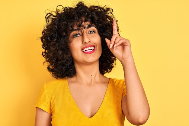 Młoda arabka z kręconymi włosami w koszulce stojąca nad odizolowanym żółtym tłem zaskoczona pomysłem lub pytaniem wskazującym palcem z radosną twarzą numer jeden