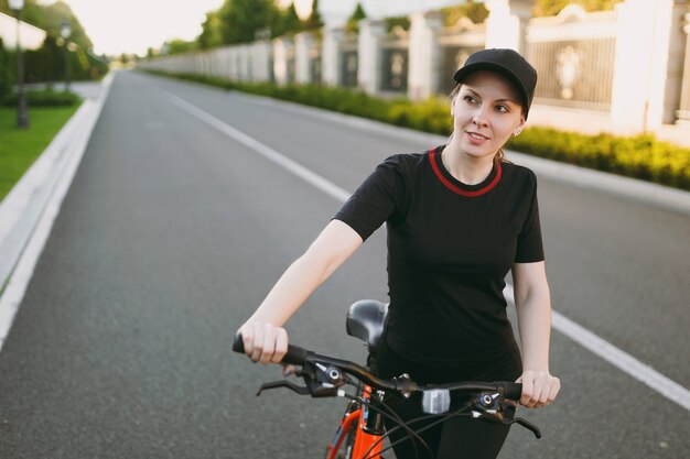 Młoda atletyczna brunetka silna kobieta w czarnym mundurze, czapka stop jazda droga na czarnym rowerze z pomarańczowymi elementami na zewnątrz na wiosenny lub letni słoneczny dzień. fitness, sport, koncepcja zdrowego stylu życia.