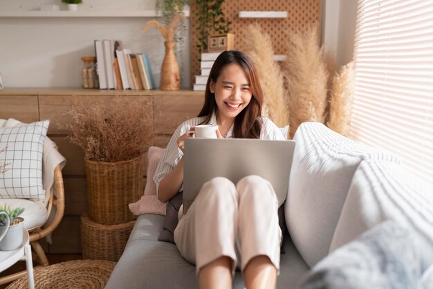 Młoda atrakcyjna azjatycka kobieta pracująca w domu ręka używa laptopa sprawdzanie harmonogramu wyszukiwania raport planowania procesu pracy z wypoczynkiem relaks na kanapie w salonie z porannym światłem spokojnym momentem
