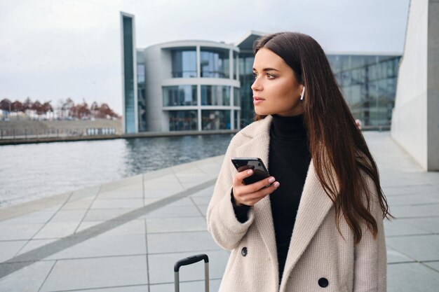 Młoda atrakcyjna bizneswoman w bezprzewodowych słuchawkach rozważnie używająca telefonu komórkowego na zewnątrz