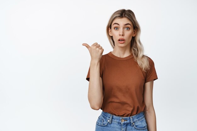 Młoda atrakcyjna dziewczyna ze zdziwioną twarzą wyglądającą na zaciekawioną i zadającą pytanie wskazującym palcem w lewo na banerze z logo pokazującym białe tło reklamy