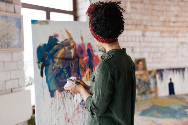 Młoda atrakcyjna kobieta o ciemnych kręconych włosach od tyłu marzycielsko rysuje obraz na płótnie ręcznie jasnymi farbami olejnymi, spędzając czas w dużym przytulnym warsztacie