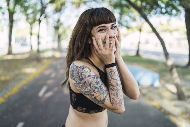 Młoda atrakcyjna kobieta rasy kaukaskiej z tatuażami stoi w parku i robi śliczną minę