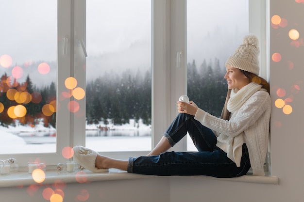 Młoda atrakcyjna kobieta w stylowym białym swetrze z dzianiny, szaliku i czapce siedzi w domu na parapecie na boże narodzenie trzymając szklaną kulę śnieżną obecną dekorację, zimowy widok na las, światła bokeh