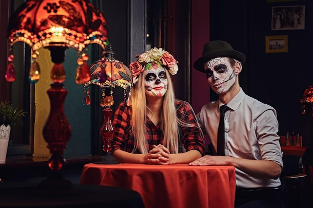 Młoda atrakcyjna para z nieumarłym makijażem czeka na zamówienie podczas randki w meksykańskiej restauracji. Koncepcja Halloween i Muertos.