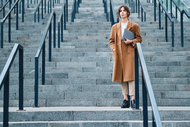 Młoda atrakcyjna przypadkowa bizneswoman w płaszczu z laptopem w zamyśleniu odwracająca wzrok na zewnątrz