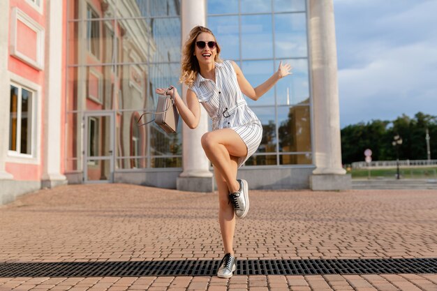 Młoda atrakcyjna stylowa kobieta bieganie, skoki śmieszne w trampki na ulicy miasta w białej sukni w stylu letniej mody na sobie okulary przeciwsłoneczne i torebkę