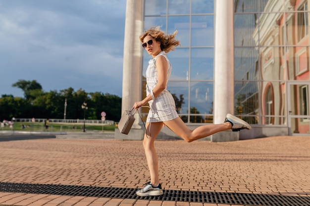 Młoda Atrakcyjna Stylowa Kobieta Bieganie, Skoki śmieszne W Trampki Na Ulicy Miasta W Białej Sukni W Stylu Letniej Mody Na Sobie Okulary Przeciwsłoneczne I Torebkę