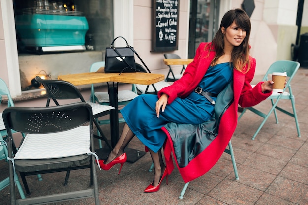 Młoda atrakcyjna stylowa kobieta siedzi w kawiarni ulicy miasta w czerwonym płaszczu picia kawy na sobie niebieską sukienkę