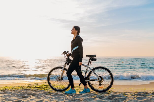 Młoda atrakcyjna szczupła kobieta jedzie na rowerze, sport na plaży o poranku wschód słońca w sportowej fitness sportowej odzieży, aktywny zdrowy tryb życia, uśmiechnięta szczęśliwa zabawa