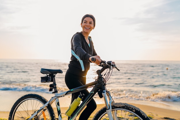 Młoda atrakcyjna szczupła kobieta jedzie na rowerze, sport na plaży o poranku wschód słońca w sportowej fitness sportowej odzieży, aktywny zdrowy tryb życia, uśmiechnięta szczęśliwa zabawa