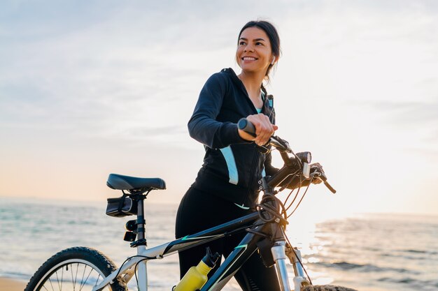 Młoda atrakcyjna szczupła kobieta jedzie na rowerze, sport w porannym wschodzie słońca letniej plaży w sportowej odzieży fitness, aktywny zdrowy tryb życia, uśmiechnięta szczęśliwa zabawa