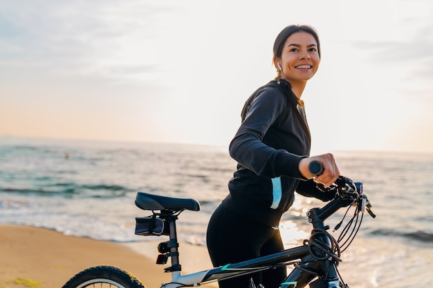 Młoda atrakcyjna szczupła kobieta jedzie na rowerze, sport w porannym wschodzie słońca letniej plaży w sportowej odzieży fitness, aktywny zdrowy tryb życia, uśmiechnięta szczęśliwa zabawa