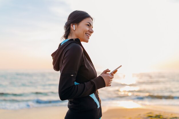 Młoda atrakcyjna szczupła kobieta robi ćwiczenia sportowe na plaży o poranku wschód słońca w strojach sportowych, zdrowy styl życia, słuchanie muzyki na słuchawkach bezprzewodowych trzymając smartfon, uśmiechając się szczęśliwy, bawiąc się