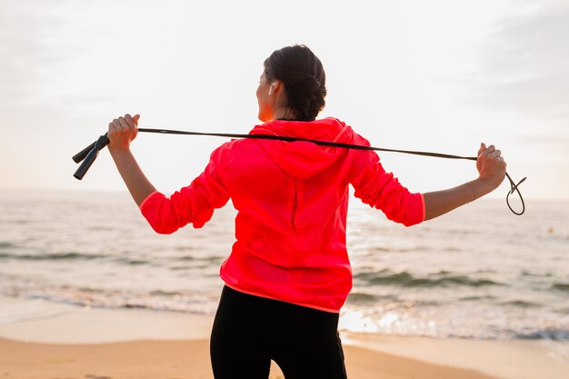 Młoda atrakcyjna szczupła kobieta robi ćwiczenia sportowe o porannym wschodzie słońca na plaży w odzieży sportowej, zdrowym stylu życia, słuchaniu muzyki na słuchawkach, na sobie różową wiatrówkę, trzymając skakankę