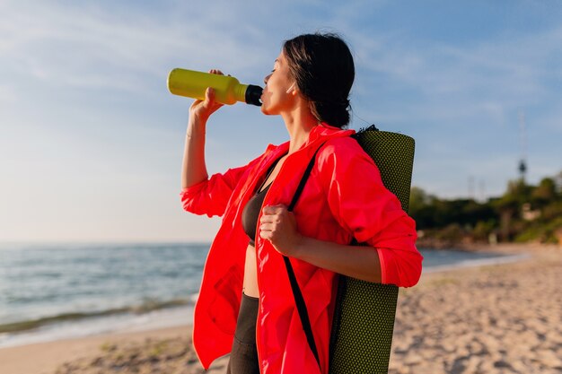 Młoda atrakcyjna uśmiechnięta kobieta uprawia sport o porannym wschodzie słońca na plaży morskiej trzymając matę do jogi i butelkę wody, zdrowy styl życia, słuchanie muzyki na słuchawkach, na sobie różową wiatrówkę