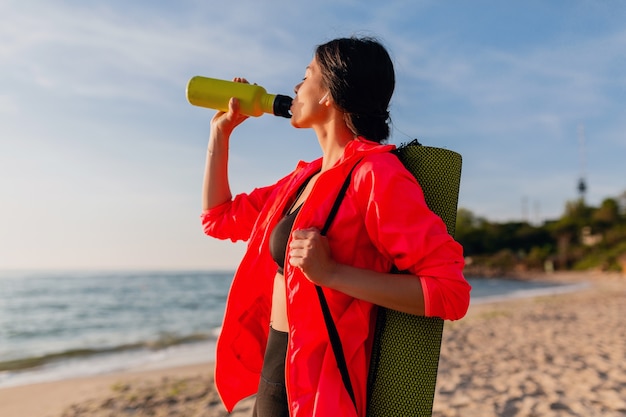 Młoda atrakcyjna uśmiechnięta kobieta uprawia sport o porannym wschodzie słońca na plaży morskiej trzymając matę do jogi i butelkę wody, zdrowy styl życia, słuchanie muzyki na słuchawkach, na sobie różową wiatrówkę