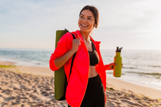 Młoda Atrakcyjna Uśmiechnięta Kobieta Uprawia Sport O Porannym Wschodzie Słońca Na Plaży Morskiej Trzymając Matę Do Jogi I Butelkę Wody, Zdrowy Styl życia, Słuchanie Muzyki Na Słuchawkach, Na Sobie Różową Wiatrówkę