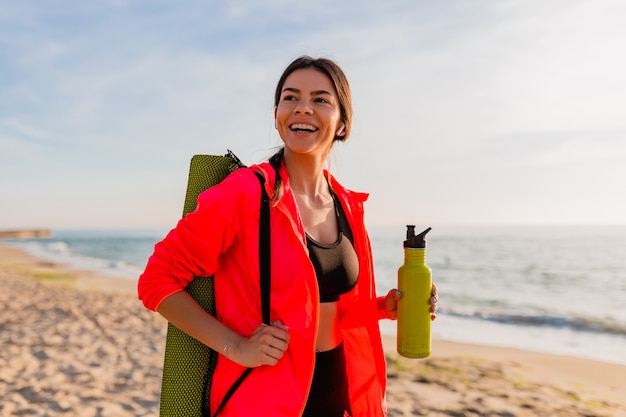 Młoda atrakcyjna uśmiechnięta kobieta uprawia sport o porannym wschodzie słońca na plaży morskiej trzymając matę do jogi i butelkę wody, zdrowy styl życia, słuchanie muzyki na słuchawkach, na sobie różową wiatrówkę
