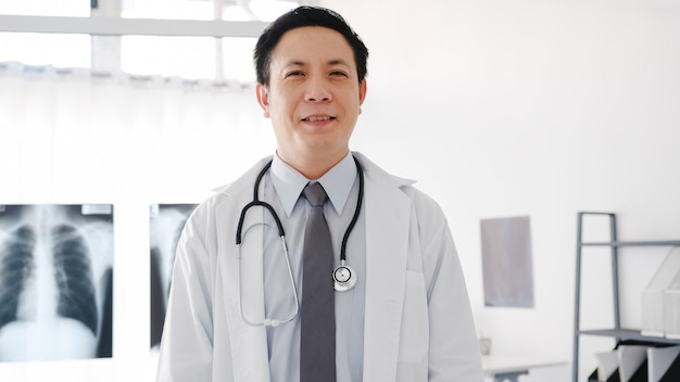 Młoda Azja mężczyzna lekarz w białym mundurze medycznym ze stetoskopem, patrząc na kamerę, uśmiech i ramiona skrzyżowane podczas wideokonferencji z pacjentem w szpitalu.