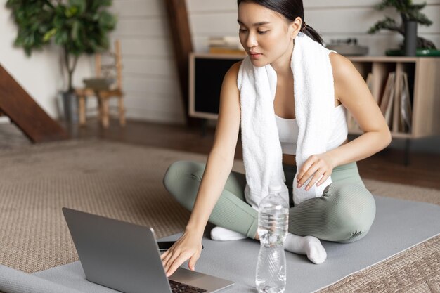 Młoda Azjatka łączy się z instruktorem fitness online na laptopie, ćwicząc za pomocą czatu wideo w domu...