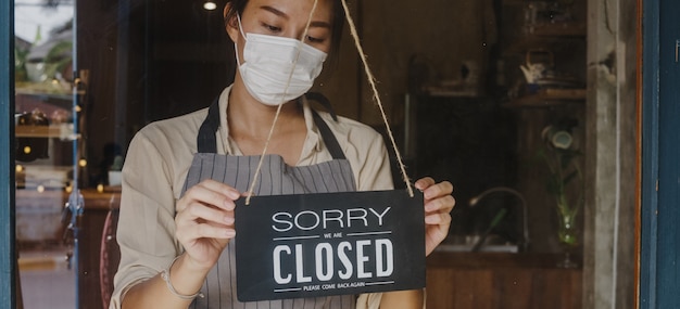 Młoda Azjatka nosi maskę na twarz, zmieniając znak z otwartego na zamknięty w kawiarni ze szklanymi drzwiami po kwarantannie koronawirusa.