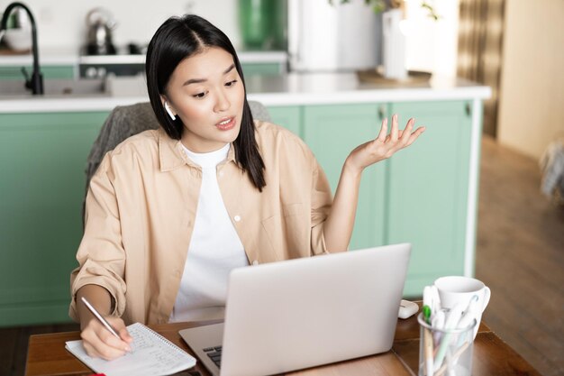 Młoda Azjatka pracująca zdalnie z domu, zapisująca notatki, studentka studiująca wykład online...