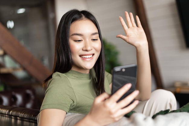 Młoda Azjatka wideorozmawia przez telefon komórkowy macha ręką przed kamerą smartfona rozmawiając na komórce...