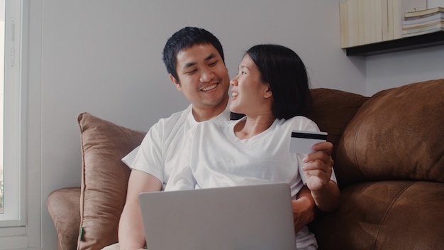 Młoda Azjatycka Ciężarna para robi zakupy online w domu. Mama i tata czują się szczęśliwi, używając technologii laptopa i karty kredytowej kupując produkt dla dziecka, leżąc na kanapie w salonie w domu.
