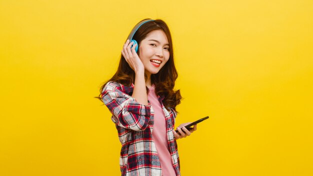 Młoda Azjatycka dama jest ubranym bezprzewodowych hełmofony słucha muzyka od smartphone z rozochoconym wyrażeniem w przypadkowej odzieży i patrzeje kamerę nad kolor żółty ścianą. Koncepcja wyrazu twarzy.