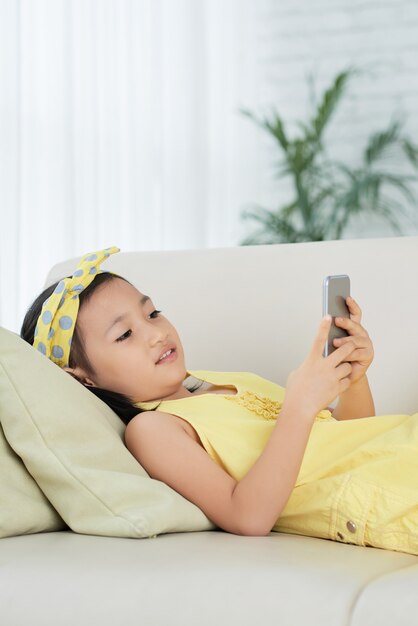 Młoda Azjatycka dziewczyna kłama na kanapie w domu i używa smartphone