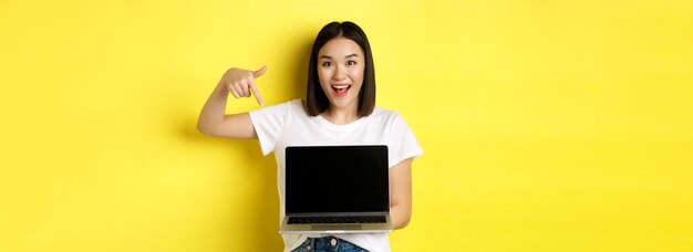 Młoda azjatycka kobieta demonstruje ofertę online pokazującą ekran laptopa i uśmiechniętą pozycję nad żółtymi półdupkami