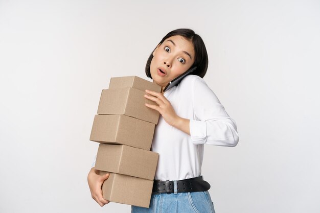 Młoda azjatycka kobieta odpowiada na telefon, rozmawia przez telefon komórkowy, niosąc stos pudełek z zamówieniami stojącymi na białym tle