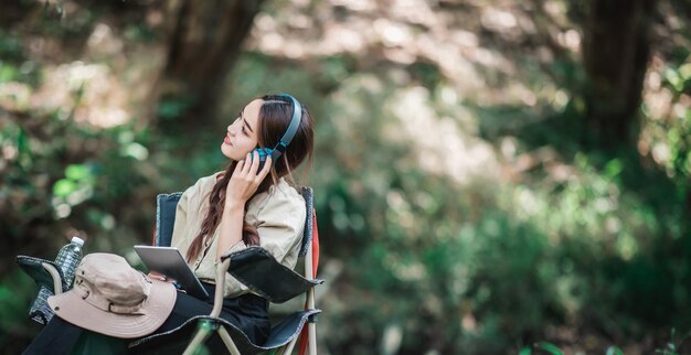 Młoda azjatycka kobieta siedzi na krześle w pobliżu strumienia, słuchając muzyki z tabletu za pomocą bezprzewodowych słuchawek podczas biwakowania w lesie