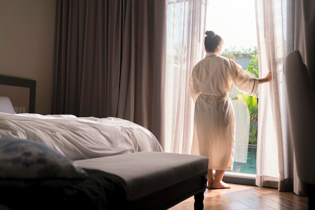 Młoda azjatycka kobieta stoi otwarte białe zasłony przy oknie rano po przebudzeniu w hotelowej sypialni Kobieta budzi się ze świeżymi i otwiera zasłony w oknach