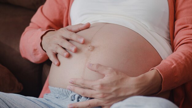 Młoda Azjatycka kobieta w ciąży trzyma jej brzuch opowiada z jej dzieckiem. Mama czuje się szczęśliwy uśmiechnięty pozytywny i spokojny, podczas gdy dbać o dziecko, ciąża leżąc na kanapie w salonie w domu.