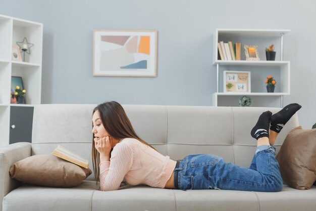 Młoda azjatycka kobieta w zwykłych ubraniach leżąca na kanapie w domu wnętrze czytająca książkę jest zaintrygowana relaksującym spędzaniem weekendu w domu