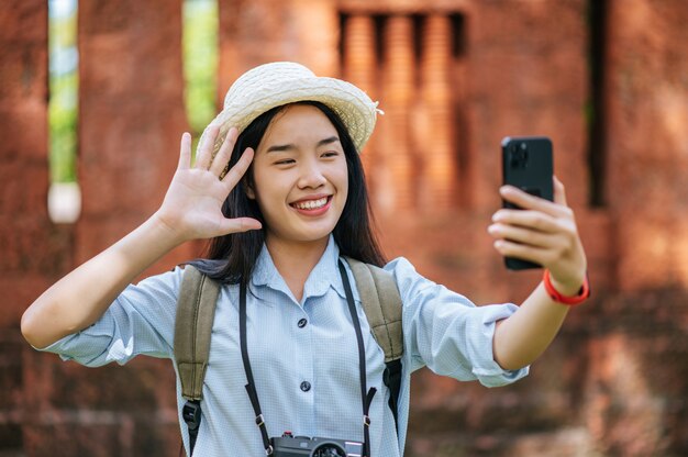 Młoda azjatycka kobieta z plecakiem w kapeluszu podróżująca po zabytkowym miejscu, używa smartfona i aparatu, robi zdjęcie z happy