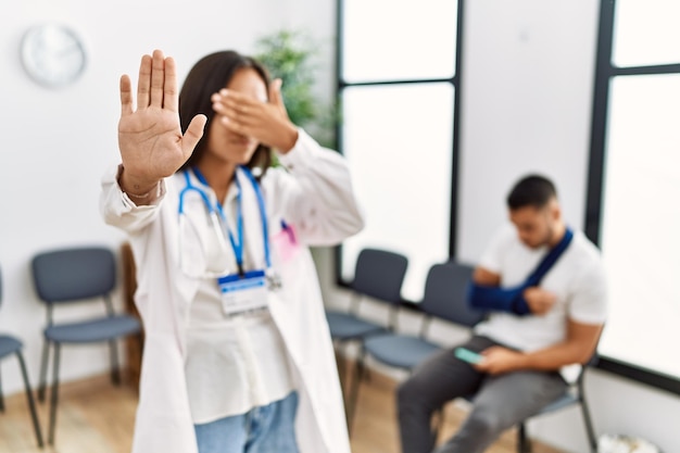 Bezpłatne zdjęcie młoda azjatycka lekarka w poczekalni z mężczyzną ze złamaną ręką zakrywającą oczy dłońmi i wykonującą gest zatrzymania z wyrazem smutku i strachu. zawstydzona i negatywna koncepcja.