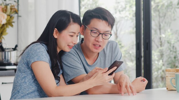 Młoda Azjatycka para cieszy się robić zakupy online na telefonie komórkowym w domu. Styl życia młody mąż i żona szczęśliwi kupują e-commerce po śniadaniu w nowoczesnej kuchni w domu rano.