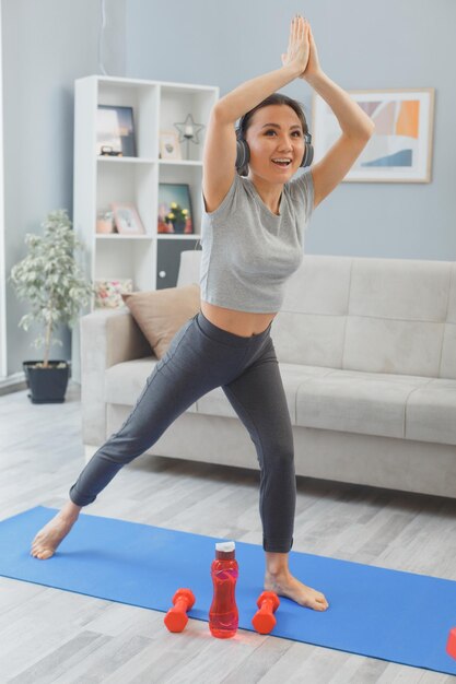 młoda azjatycka zdrowa kobieta ze słuchawkami ćwicząca w domu z butelką wody i hantlami, wykonująca ćwiczenia rozciągające na macie do jogi