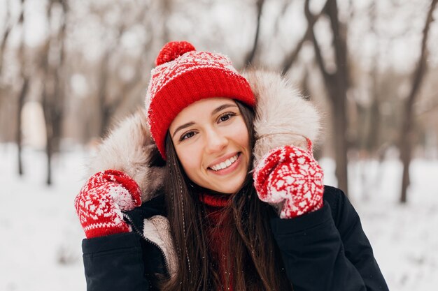 Młoda, bardzo uśmiechnięta szczęśliwa kobieta w czerwonych rękawiczkach i czapce z dzianiny w zimowym płaszczu z futrzanym kapturem, spacerująca po parku w śniegu, ciepłe ubrania