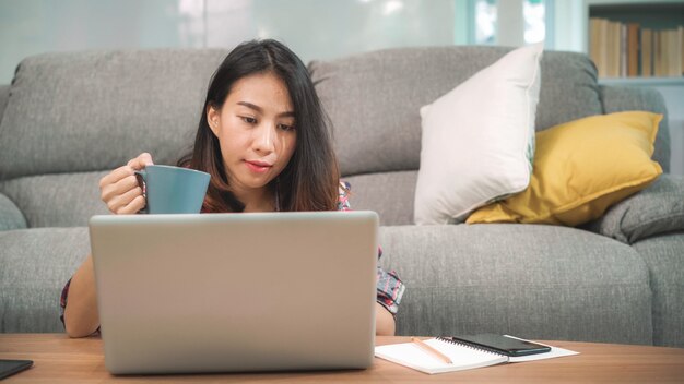 Młoda biznesowa niezależna Azjatycka kobieta pracuje na laptopie sprawdza ogólnospołecznych środki i pije kawę podczas gdy kłamający na kanapie gdy relaksuje w żywym pokoju w domu.